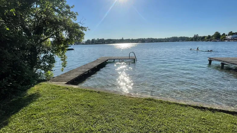 Idyllischer Seeblick mit Holzsteg und glitzerndem Wasser unter strahlender Sonne.