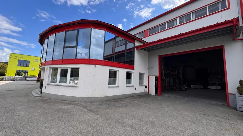 Modernes Geschäftsgebäude mit großen Glasflächen und rotem Akzent, inklusive Einfahrt zum Lagerbereich.