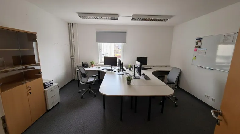 Geräumiges Büro mit mehreren Arbeitsplätzen, Teppichboden und einem Fenster mit Rollo.