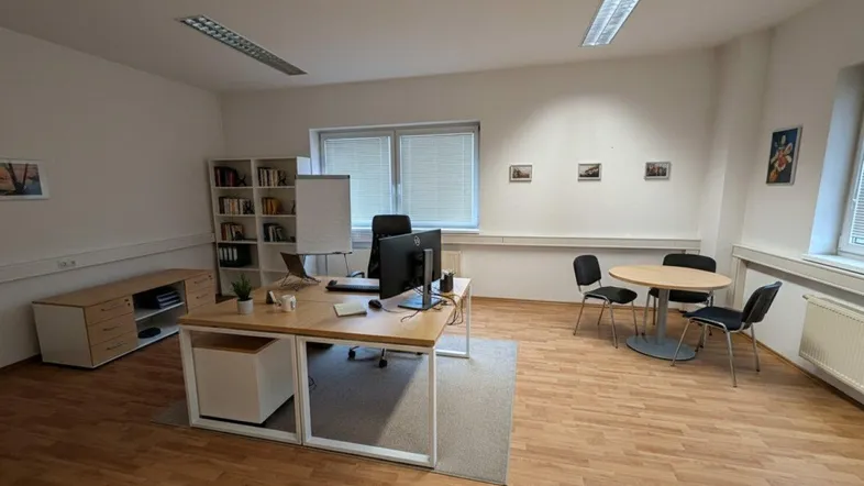 Helles Büro mit modernem Schreibtisch, Besprechungsbereich und Laminatboden, gut beleuchtet durch große Fenster.