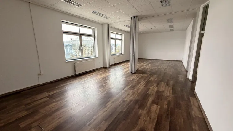 Geräumiges Büro mit dunklem Laminatboden, weißen Wänden und großen Fenstern, die viel Tageslicht hereinlassen.