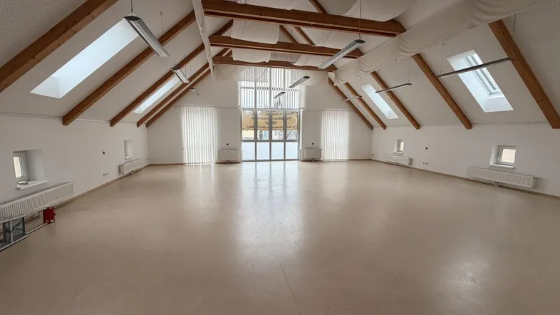 Großer, heller Mehrzweckraum mit sichtbaren Holzbalken, Dachfenstern und hellem Bodenbelag, ideal für Veranstaltungen.