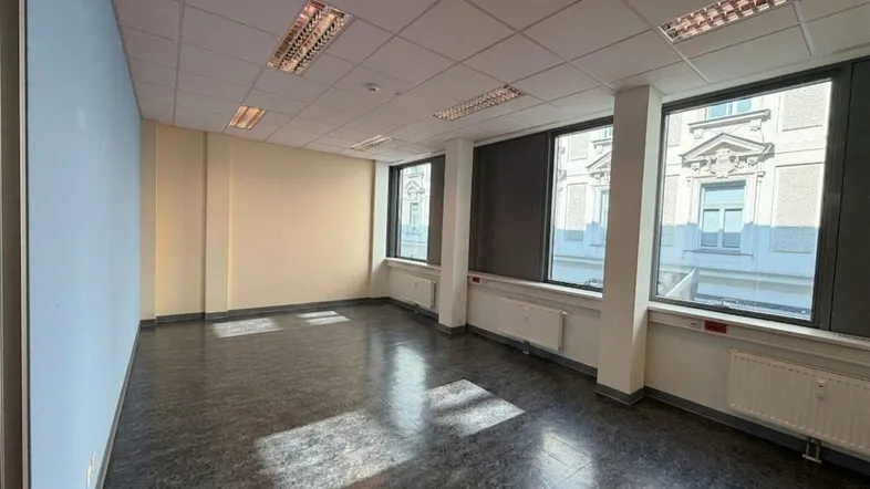 Geräumiges, leeres Büro mit großen Fenstern, die viel Tageslicht hereinlassen und einen Blick auf die Stadt bieten.