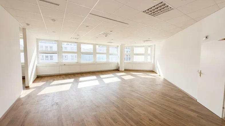 Geräumiges, leeres Büro mit großen Fenstern, Holzboden und viel natürlichem Licht.