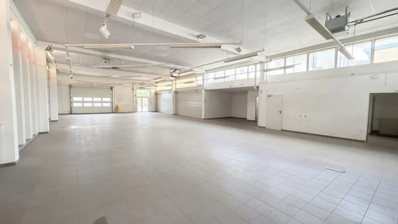Geräumige, leere Gewerbefläche mit Fliesenboden, hohen Decken und viel Tageslicht durch große Fenster.