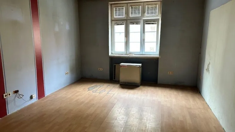 Leeres Zimmer mit Holzboden, grauen Wänden und einem Fenster, das auf den Innenhof blickt.