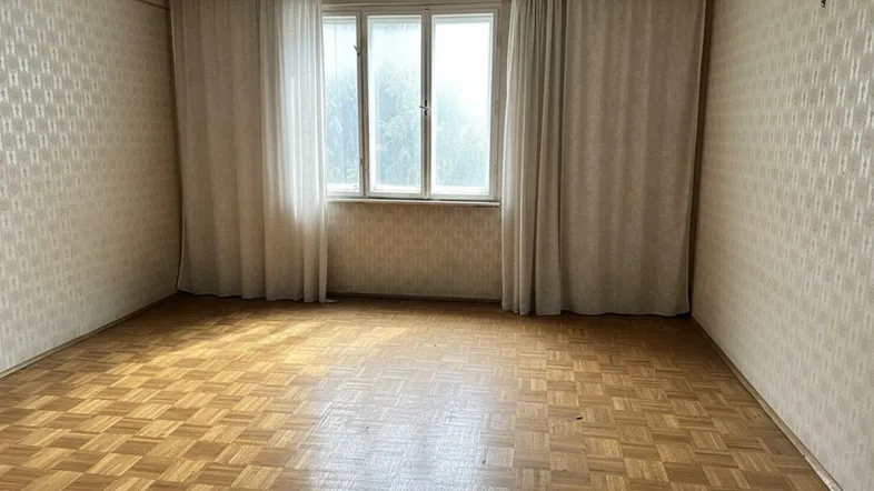 Geräumiges Zimmer mit Parkettboden, gemusterter Tapete und großem Fenster mit hellen Vorhängen.