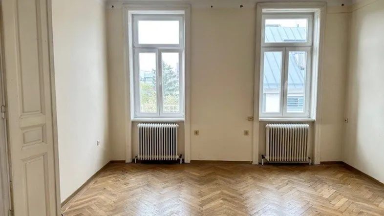 Helles Zimmer mit Fischgrätparkettboden, zwei Fenstern und Heizkörpern, bereit zur Renovierung.