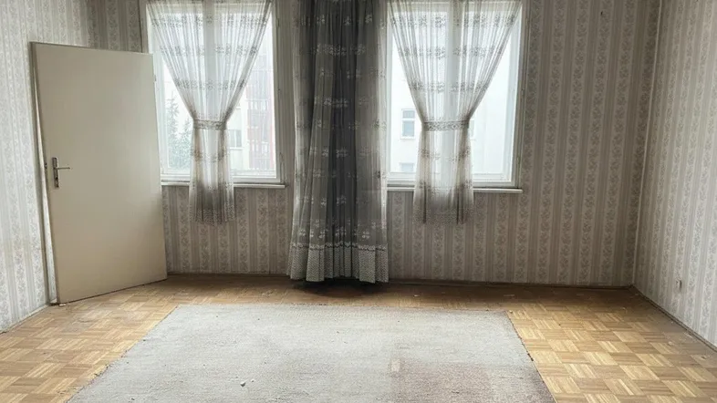 Leeres Zimmer mit gemusterter Tapete, Parkettboden und zwei Fenstern mit Vorhängen.