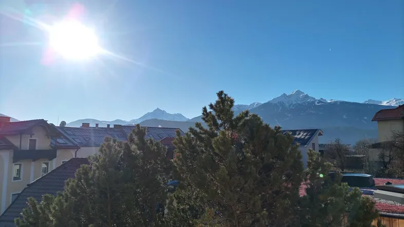 Sonniger Ausblick auf schneebedeckte Berge und umliegende Dächer mit Bäumen im Vordergrund.