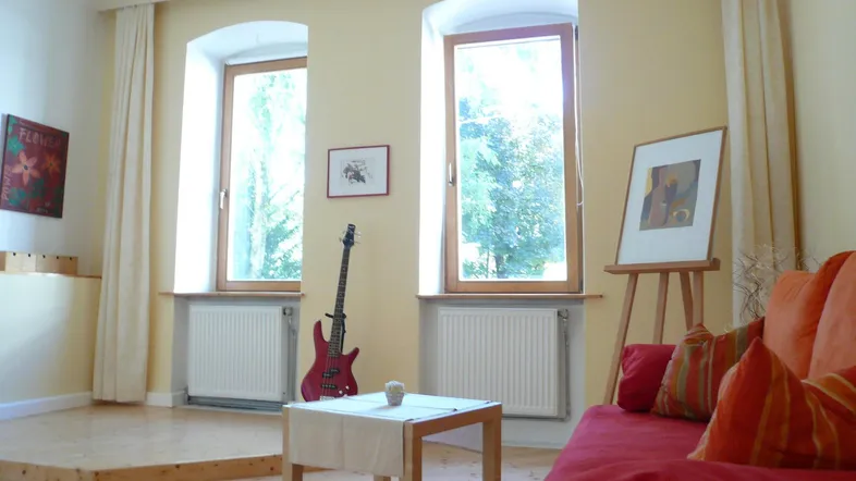 Helles Wohnzimmer mit Holzboden, rotem Sofa und zwei hohen Fenstern mit Blick ins Grüne.