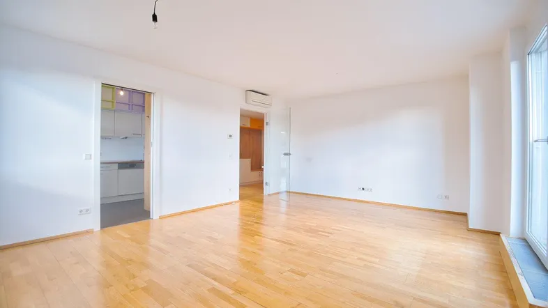 Helles, leeres Zimmer mit Parkettboden, weißen Wänden und Zugang zu Küche und Balkon.
