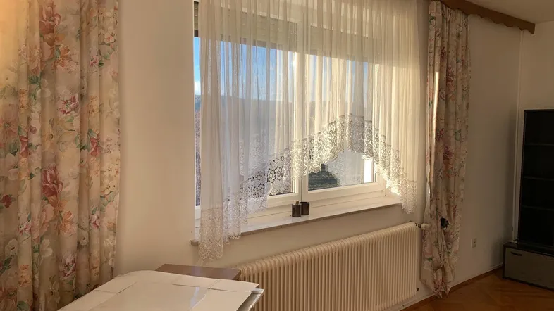 Helles Zimmer mit floralen Vorhängen, Parkettboden und einem Fenster, das viel Sonnenlicht hereinlässt.
