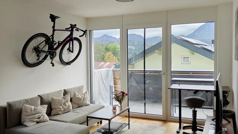 Helles Wohnzimmer mit Holzboden, Zugang zum Balkon und Bergblick durch große Fenster.