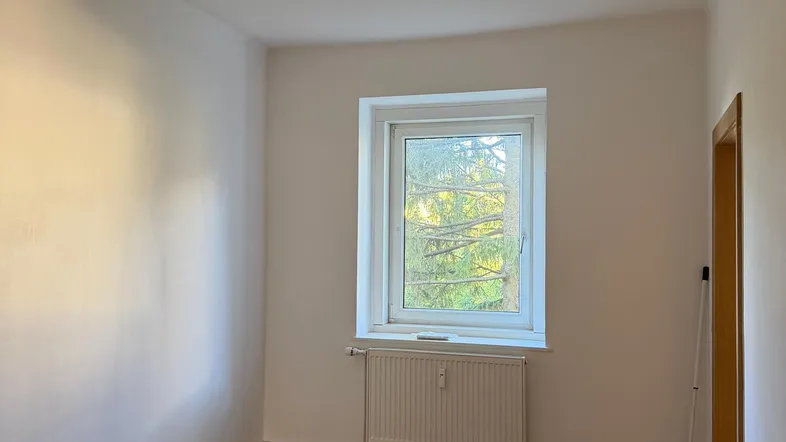 Helles, leeres Zimmer mit Laminatboden und einem Fenster, das viel Tageslicht hereinlässt.