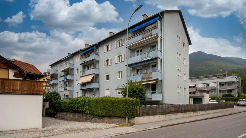 Mehrfamilienhaus mit Balkonen und Blick auf die umliegenden Berge unter blauem Himmel.