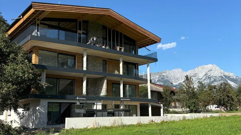 Modernes Mehrfamilienhaus mit Holzfassade und großen Balkonen vor alpiner Bergkulisse.