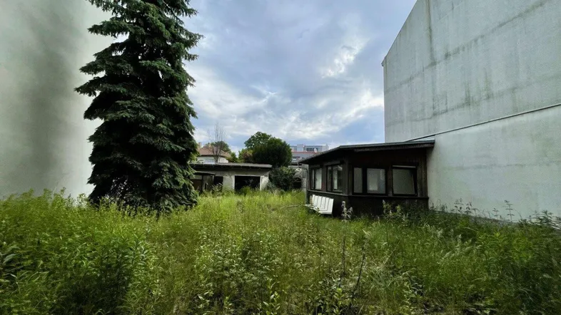 Verwildertes Grundstück mit hohem Gras, einem kleinen Nebengebäude und umgebenden Gebäudewänden unter bewölktem Himmel.