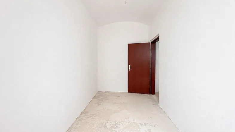 Leerer Raum mit weißen Wänden, Betonboden und einer braunen Tür, der auf eine Renovierung hindeutet.