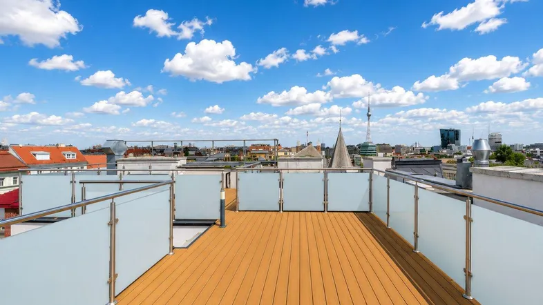 Große Dachterrasse mit Holzboden, Glasgeländer und weitem Stadtblick unter blauem Himmel.