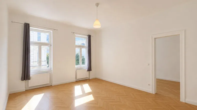 Helles Zimmer mit Fischgrätparkett und zwei großen Fenstern, das viel Tageslicht hereinlässt.