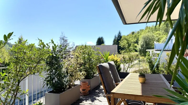 Sonnige Terrasse mit Holztisch, bequemen Stühlen und üppigen Topfpflanzen, umgeben von grüner Natur und weitem Blick.