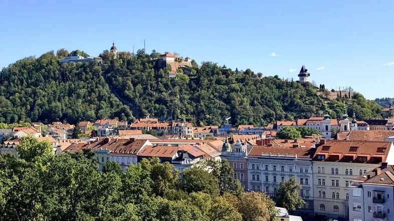 Panoramablick über die Stadt Graz mit dem bewaldeten Schlossberg unter strahlend blauem Himmel.