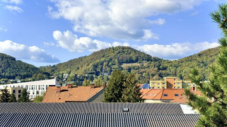 Weitblick über Dächer auf bewaldete Hügel unter einem strahlend blauen Himmel mit Wolken.