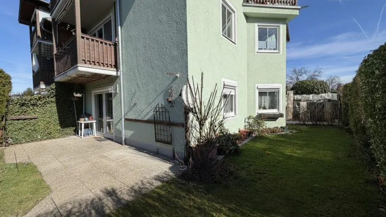 Grüne Fassade des Gebäudes mit einer gepflasterten Terrasse und angrenzendem Rasenbereich unter blauem Himmel.