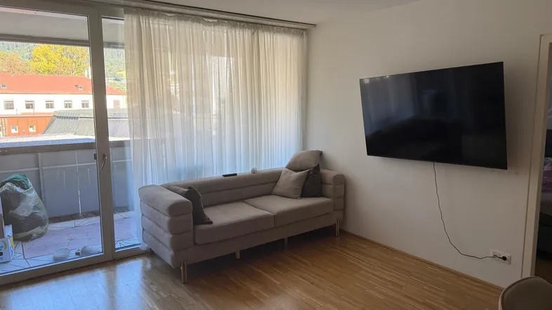 Helles Wohnzimmer mit beigem Sofa, Wandfernseher und Zugang zu einem Balkon mit Blick ins Grüne.