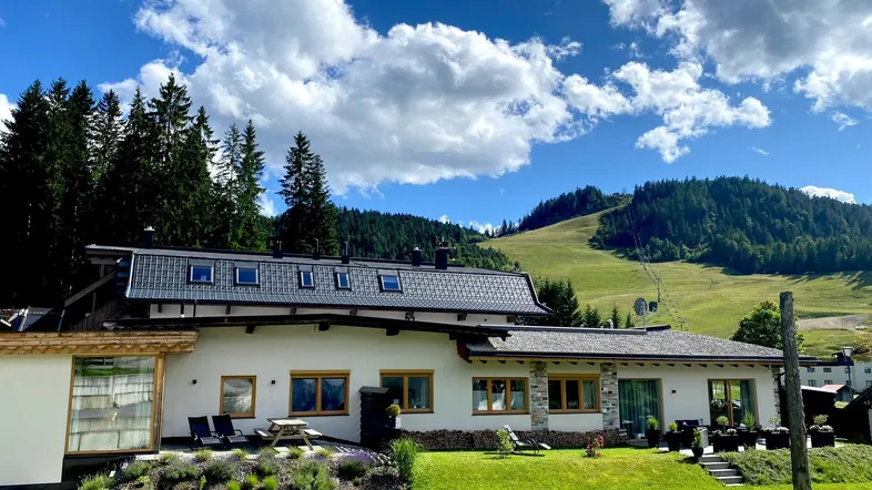 Modernes Haus mit Satteldach, umgeben von grünen Wiesen und bewaldeten Bergen unter blauem Himmel.