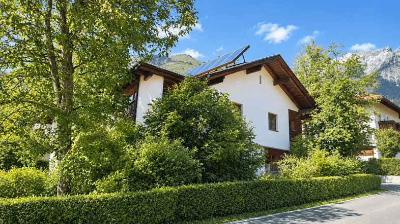 Modernes Haus mit weißer Fassade, Photovoltaikanlage und üppigem Grün in den Bergen gelegen.