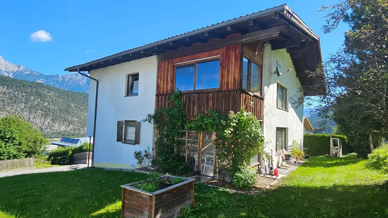 Traditionelles Haus mit Holzbalkon und begrüntem Gartenbereich vor alpiner Bergkulisse.