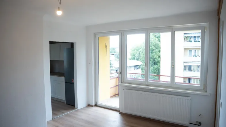 Helles Zimmer mit Laminatboden, großem Fenster und Zugang zu einem Balkon, mit Blick in die Küche.