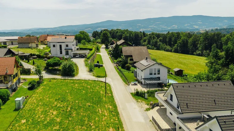 Luftaufnahme einer ländlichen Wohnsiedlung mit Häusern, grünen Feldern und bewaldeten Hügeln im Hintergrund.