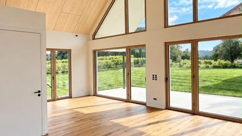 Helles Wohnzimmer mit Holzboden, hohen Decken und großen Fenstern mit Blick ins Grüne.