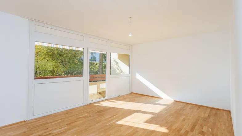 Helles, leeres Wohnzimmer mit Parkettboden und Zugang zu einem sonnigen Balkon.
