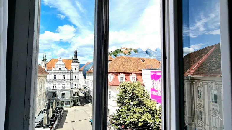 Blick aus einem Fenster auf eine belebte Stadtstraße mit historischen Gebäuden und blauem Himmel.