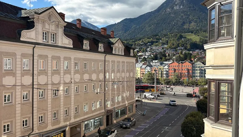 Blick von einem Balkon auf eine belebte Straße mit historischen Gebäuden und majestätischen Bergen im Hintergrund.