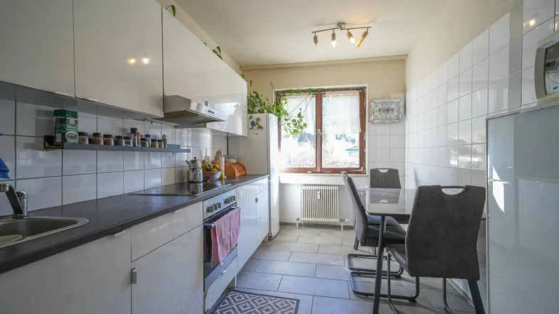 Moderne Küche mit weißen Schränken, dunkler Arbeitsplatte, Fliesenboden und Essbereich am Fenster.