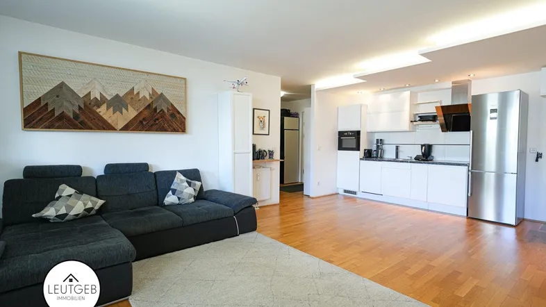 Geräumiger offener Wohnbereich mit moderner Küche, dunklem Sofa und Holzboden.