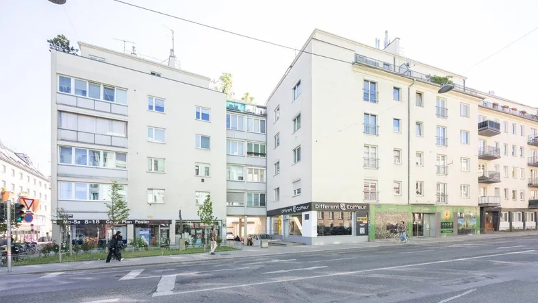 Ansicht eines Mehrfamilienhauses mit Geschäften im Erdgeschoss an einer belebten Stadtstraße.