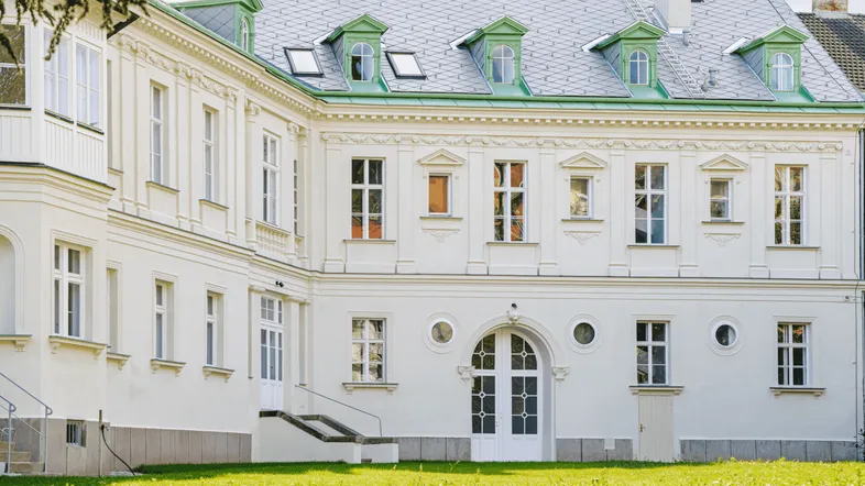 Klassische weiße Fassade eines historischen Gebäudes mit grünen Dachgauben und gepflegtem Rasen.