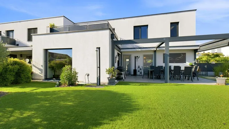 Moderne Hausfassade mit großer überdachter Terrasse, Essbereich im Freien und gepflegtem Rasen.