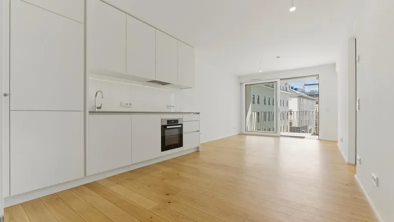 Moderne, offene Küche mit weißen Schränken und Holzboden, angrenzend an einen hellen Wohnbereich mit Balkonzugang.