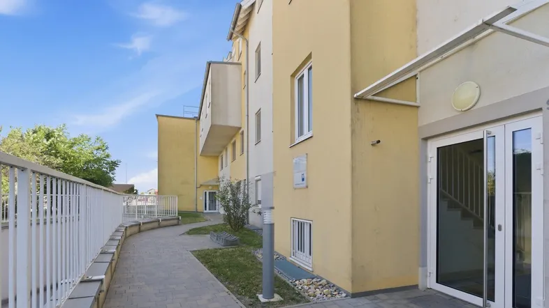 Eingangsbereich eines modernen Mehrfamilienhauses mit gepflastertem Weg und gelber Fassade unter blauem Himmel.
