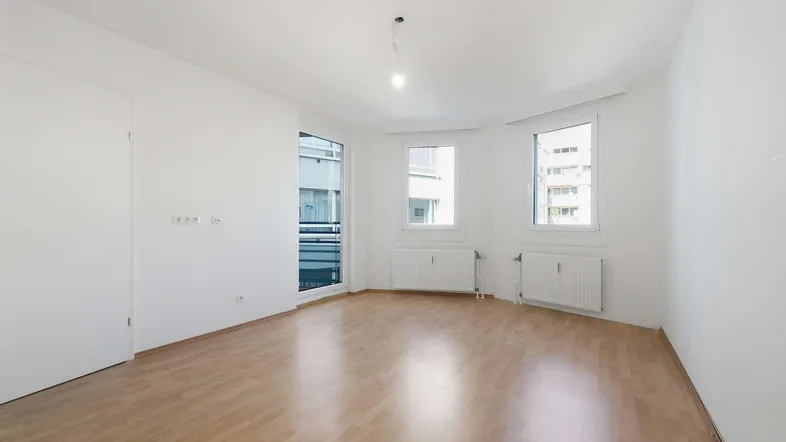 Helles, leeres Zimmer mit Parkettboden, weißen Wänden und Zugang zu einem Balkon.