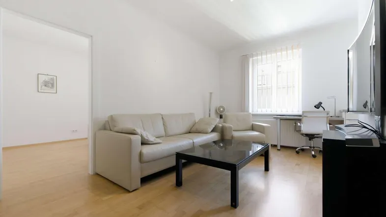 Helles Wohnzimmer mit beigem Sofa, Couchtisch und einem Arbeitsbereich am Fenster.