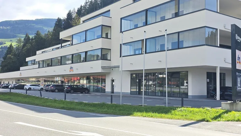 Modernes Gebäude mit großen Glasflächen, Parkplätzen und Geschäften im Erdgeschoss, umgeben von grünen Bergen.