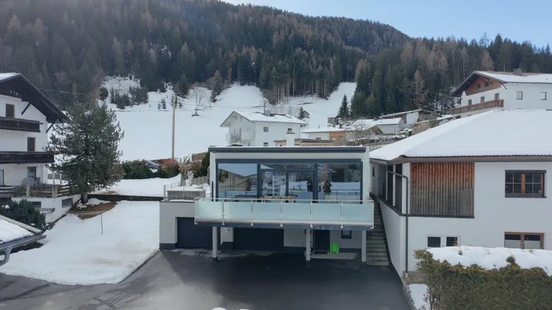 Modernes Gebäude mit großen Glasflächen und Balkon in verschneiter Berglandschaft.
